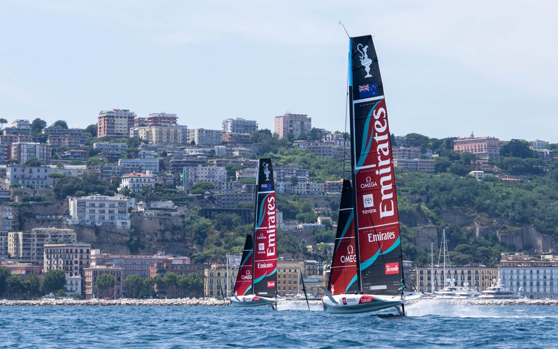 Two Emirates Team New Zealand AC40 foiling monohulls sailing in front of the Naples waterfront.