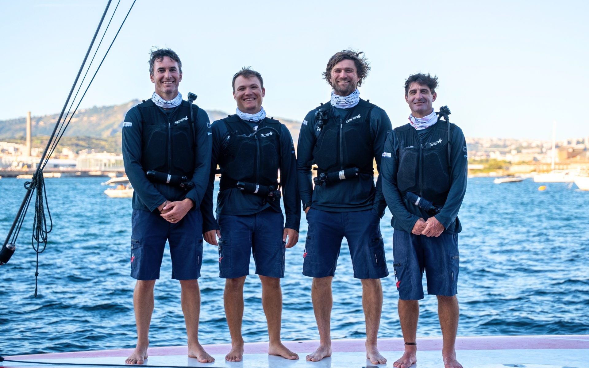 Four ARC Team USA sailors stand on deck in sailing gear with the water behind them.