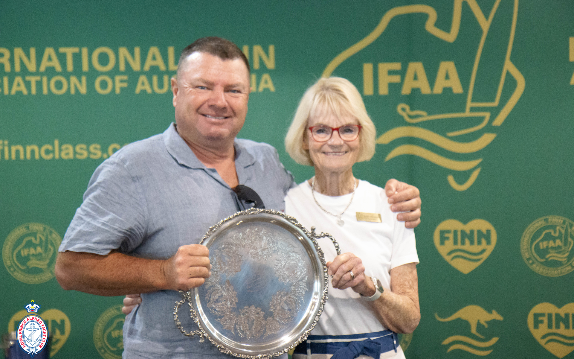 Anthony Nossiter holds a large silver trophy alongside a woman in front of a Finn association backdrop.